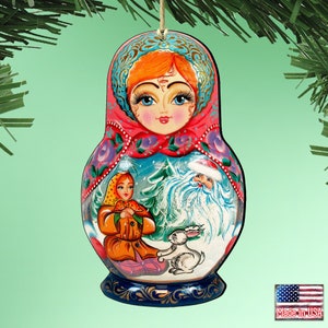 May include: A hand-painted wooden Matryoshka doll ornament. The ornament features a woman's face at the top, with a red headscarf and blue eyes. The doll's body depicts a winter scene with a woman, Santa Claus, and a rabbit. The ornament is suspended by a string.