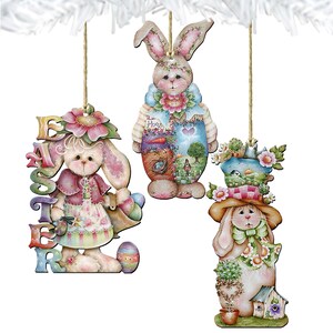 May include: Three wooden Easter bunny ornaments with floral and pastel designs. One bunny holds a basket of eggs, another has a painted scene, and the third wears a hat and scarf. Each ornament is suspended by a natural twine string.