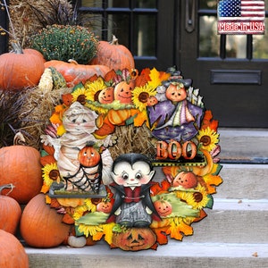 Peut inclure: Une couronne d'Halloween avec des personnages de momie, de vampire et de sorci&egrave;re. La couronne est d&eacute;cor&eacute;e de tournesols, de citrouilles et de feuilles d'automne. Le texte "BOO" est affich&eacute; sur la couronne.