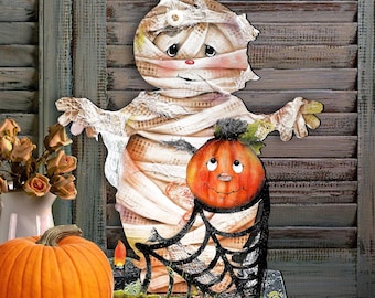 May include: A decorative wooden cutout featuring a mummy character wrapped in beige bandages with a small white button detail, holding an orange pumpkin with a black spiderweb base. The mummy has a painted face with large eyes and a small nose.