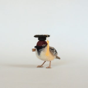 May include: A small, brown and white bird wearing a black graduation cap with a yellow tassel. The bird is holding a small, brown stick in its beak.