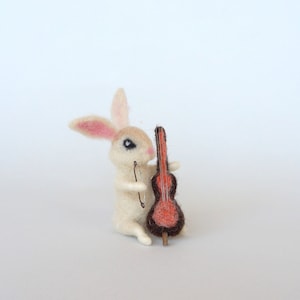 May include: A white felt rabbit figurine playing a brown felt cello with a red body. The rabbit has pink ears and black eyes.