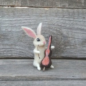 May include: A white felt rabbit holding a small red and brown felt cello. The rabbit has black eyes and pink ears.