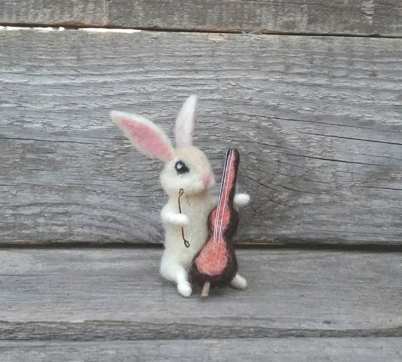 May include: A white felt rabbit holding a small red and brown felt cello. The rabbit has black eyes and pink ears.