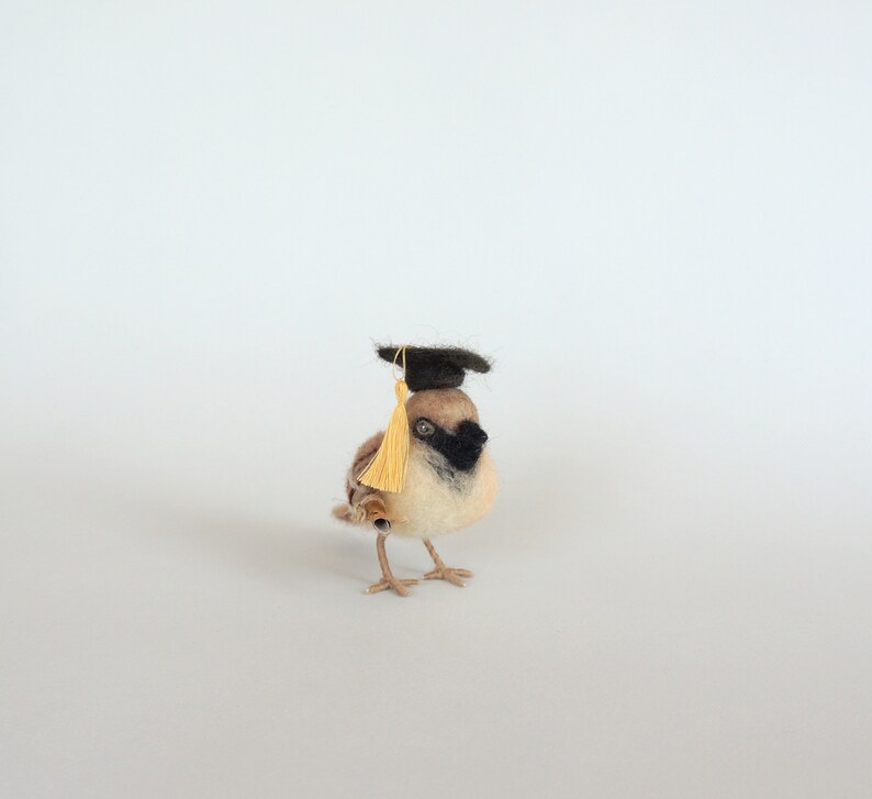 May include: A small, brown and white felt bird wearing a black graduation cap with a gold tassel. The bird is standing on a white surface.