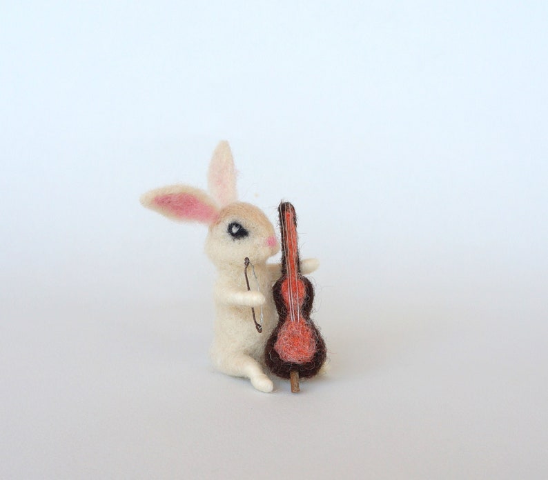 May include: A white felt rabbit figurine playing a brown felt cello with a red body. The rabbit has pink ears and black eyes.
