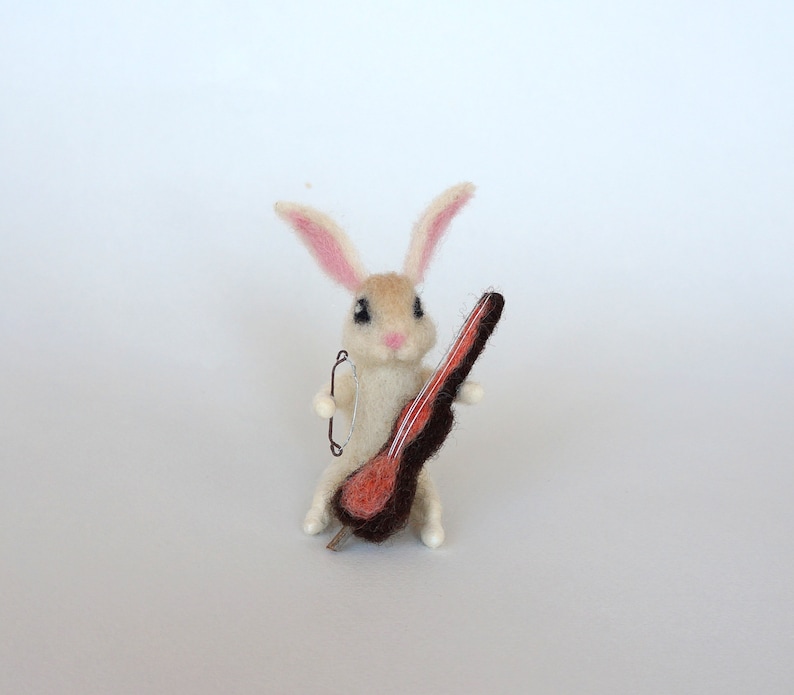 May include: A white felt rabbit with pink ears holding a small red and brown felt cello.
