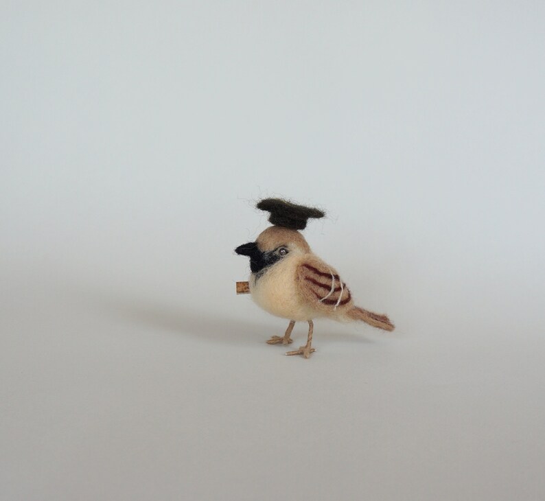 May include: A small, brown and white felt bird wearing a black felt graduation cap. The bird is standing on a white surface.