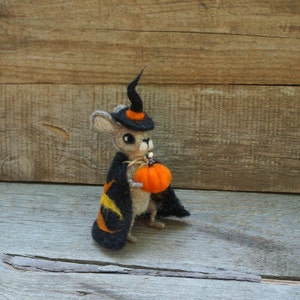 Op de afbeelding: Een kleine, grijze, vilten muis met een zwarte heksenhoed en een oranje pompoen in zijn hand. De muis staat op een houten oppervlak.