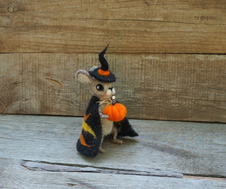 Op de afbeelding: Een kleine, grijze, vilten muis met een zwarte heksenhoed en een oranje pompoen in zijn hand. De muis staat op een houten oppervlak.
