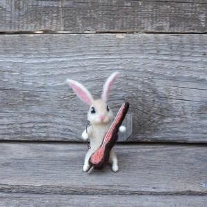 May include: A white felt rabbit holding a brown and pink felt object that resembles a guitar.