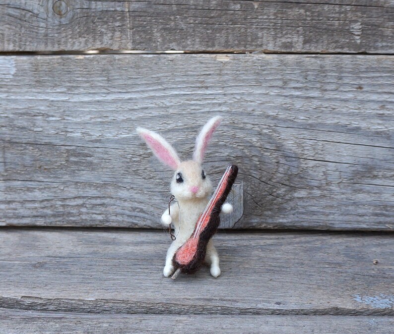 May include: A white felt rabbit holding a brown and pink felt object that resembles a guitar.