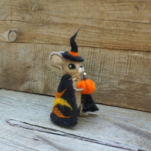 Op de afbeelding: Een kleine, vilten muis die een zwarte heksenhoed draagt met oranje en gele accenten, en een kleine oranje pompoen vasthoudt.