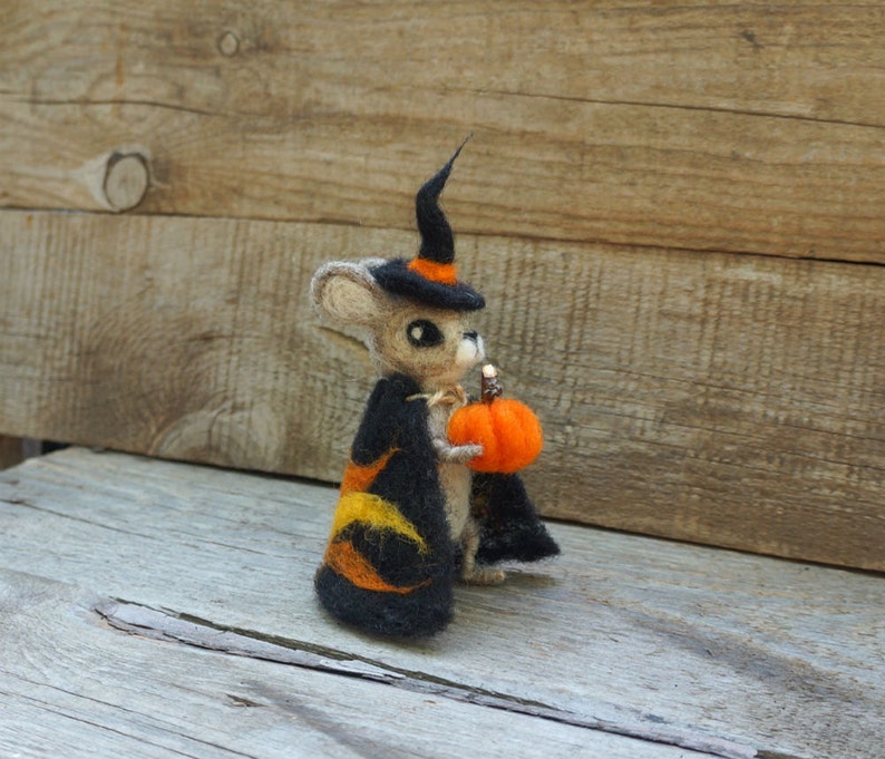 Op de afbeelding: Een kleine, vilten muis die een zwarte heksenhoed draagt met oranje en gele accenten, en een kleine oranje pompoen vasthoudt.