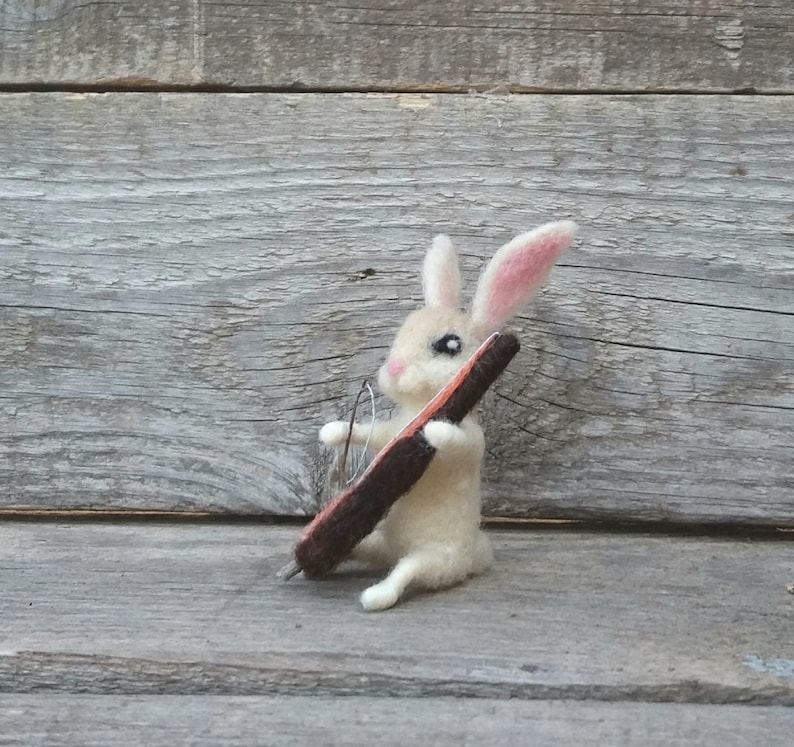 May include: A white felt rabbit with pink ears playing a brown and orange cello. The rabbit is sitting on a wooden surface.