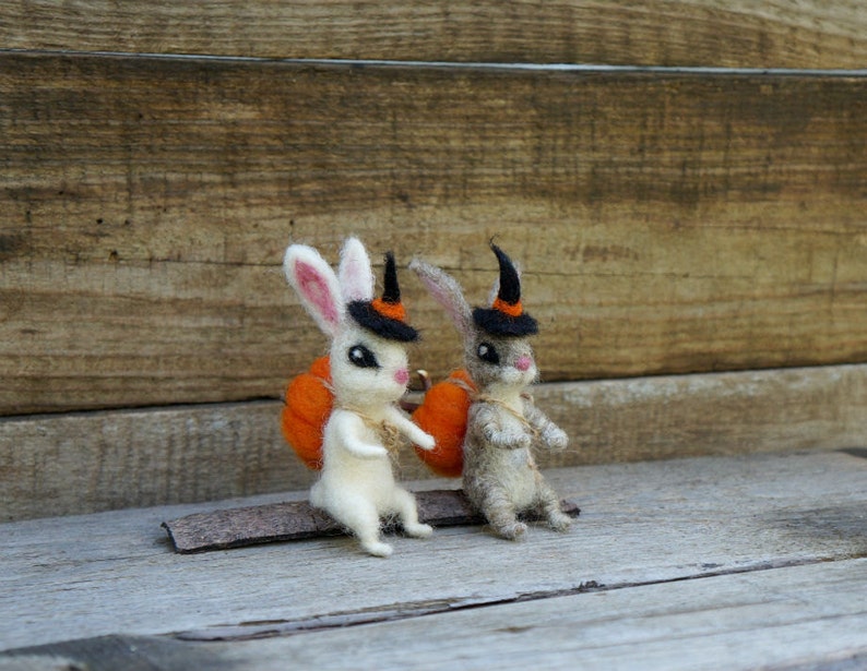 Op de afbeelding: Twee viltkonijnen die toverhoeden dragen en oranje pompoenen dragen. De konijnen zitten op een houten oppervlak.