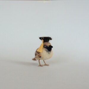 May include: A small, brown and white felt bird wearing a black graduation cap with a gold tassel. The bird is standing on a white surface.