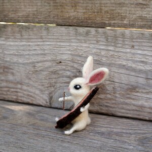 May include: A white felt rabbit with pink ears holding a brown stick. The rabbit is sitting on a wooden surface.