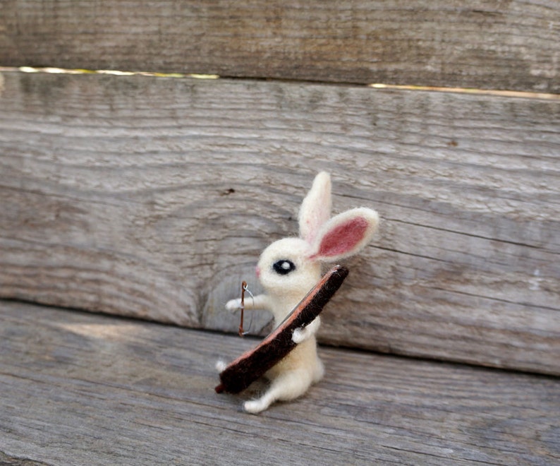 May include: A white felt rabbit with pink ears holding a brown stick. The rabbit is sitting on a wooden surface.
