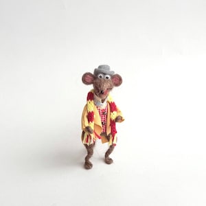 Könnte beinhalten: Eine skurrile Filzmaus-Figur, gekleidet in einem gelben Mantel, einem rot-gelben Schal und einem grauen Hut. Die Maus trägt ein rot-weiß kariertes Hemd und eine braune Hose. Die handgefertigte Maus steht auf einer weißen Oberfläche.