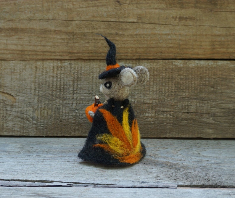 Op de afbeelding: Een viltmuis met een zwarte heksenhoed en een oranje-gele cape die een kleine pompoen vasthoudt.
