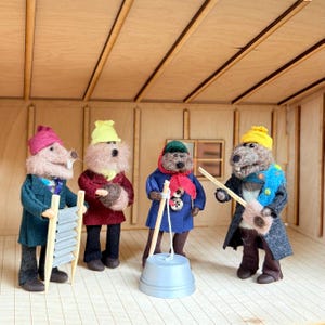 Puede incluir: Cuatro figuras de animales de lana fieltrada de pie en una habitación de madera. La primera lleva un gorro rosa y un abrigo verde azulado, sosteniendo una tabla de lavar. La segunda lleva un gorro amarillo y un abrigo granate. La tercera lleva un gorro verde y un abrigo azul con una bufanda roja. La cuarta lleva un gorro amarillo y un abrigo gris con una bufanda azul, sosteniendo un instrumento de cuerda.