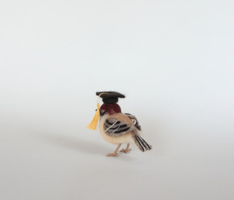 May include: A small brown and gray bird wearing a black graduation cap with a yellow tassel. The bird is standing on a white surface.