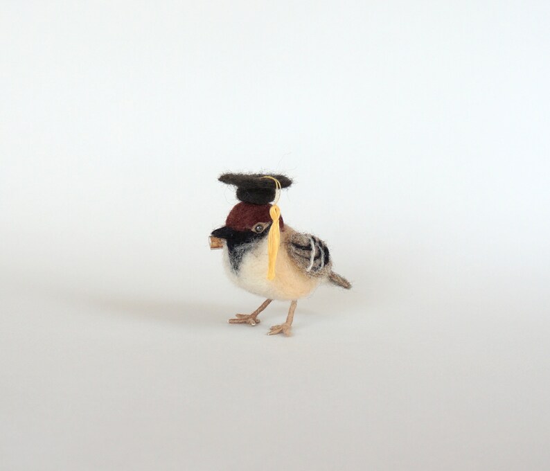 May include: A small, brown and white bird wearing a black graduation cap with a yellow tassel. The bird is made of felt and is a handmade craft.