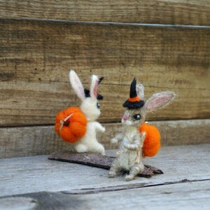Op de afbeelding: Twee vilt konijnenfiguren gekleed in Halloween-kostuums. E&eacute;n konijn draagt ​​een heksenhoed en draagt ​​een kleine oranje pompoen. Het andere konijn draagt ​​een grotere oranje pompoen.