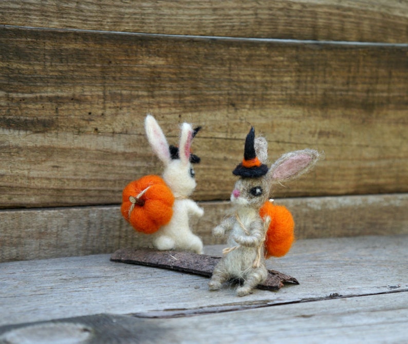 Op de afbeelding: Twee vilt konijnenfiguren gekleed in Halloween-kostuums. E&eacute;n konijn draagt ​​een heksenhoed en draagt ​​een kleine oranje pompoen. Het andere konijn draagt ​​een grotere oranje pompoen.