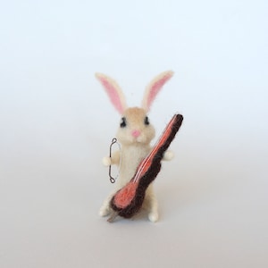 May include: A white felt rabbit with pink ears holding a brown and orange felt cello.