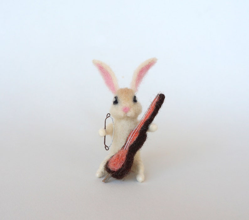 May include: A white felt rabbit with pink ears holding a brown and orange felt cello.