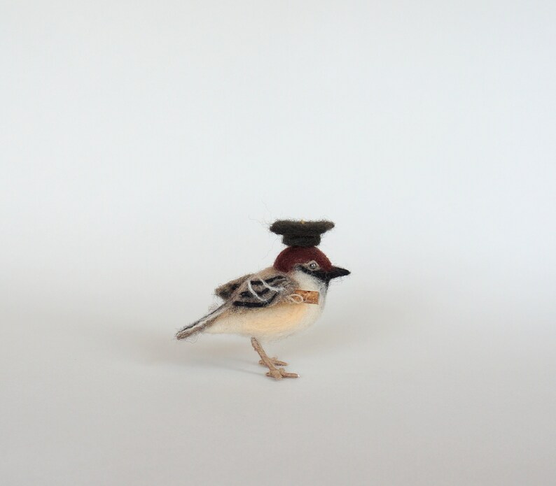 May include: A small, brown and white bird wearing a black felt hat. The bird is made of felt and has a realistic appearance.