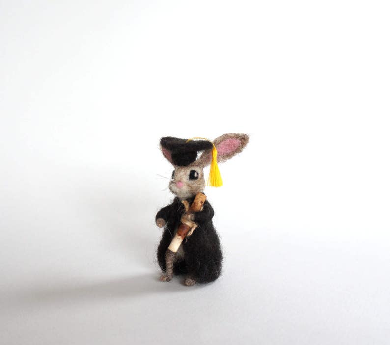 May include: A small, gray rabbit wearing a black graduation cap and gown, holding a scroll. The rabbit has yellow ears and a yellow tassel on the cap.