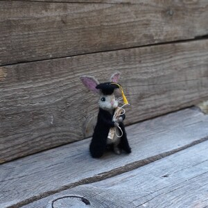May include: A small, gray felt rabbit wearing a black graduation cap and gown, holding a rolled-up scroll. The rabbit has a yellow tassel on its cap.