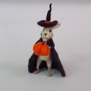 Op de afbeelding: Een wit vilt konijn met een zwarte heksenhoed en een zwarte cape, die een oranje viltpompoen vasthoudt.