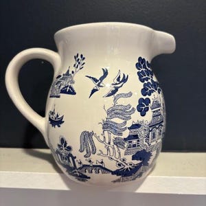 May include: White ceramic pitcher with a blue Willow pattern design. The pitcher features a curved handle and a spout. The blue design includes pagodas, birds, and other traditional elements. A classic piece for serving beverages.