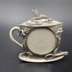 May include: A silver-toned metal picture frame shaped like a teacup on a saucer. The frame features a circular opening, a handle, and a lid topped with a sculpted hummingbird and floral details. A small spoon is attached to the saucer base.