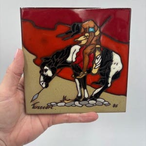 May include: Square ceramic tile with a Native American equestrian scene. The artwork features a rider on a black and white horse against a red and tan background. The tile is held by a hand.