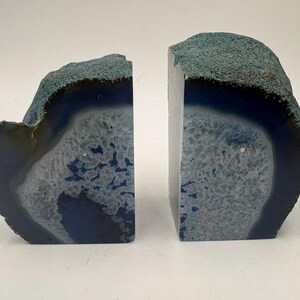 May include: A pair of bookends crafted from natural agate stone. Each piece features a polished, flat front face displaying concentric bands of deep blue and white crystalline patterns, while the outer edges retain a rough, textured blue crust.
