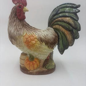 May include: A ceramic rooster figurine featuring a red comb and wattle, a yellow beak, and plumage in shades of brown, tan, and orange. The rooster stands on a base next to a small orange pumpkin and a wooden barrel, with a large, dark green tail.