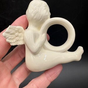 May include: A cream-colored ceramic angel napkin ring. The angel is depicted in a seated position with one wing visible. The napkin ring is a circular design held by the angel's hands. The angel has a detailed hairstyle and is resting on a black surface.
