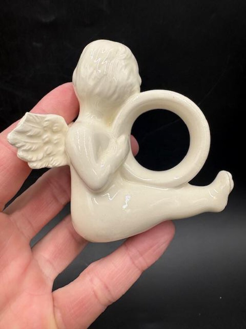 May include: A cream-colored ceramic angel napkin ring. The angel is depicted in a seated position with one wing visible. The napkin ring is a circular design held by the angel's hands. The angel has a detailed hairstyle and is resting on a black surface.