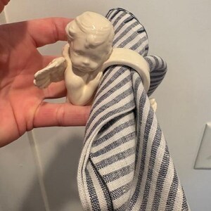 May include: A cream-colored ceramic napkin ring featuring a cherub with small wings. The cherub holds a blue and white striped napkin. The napkin ring is a decorative item for the dining table.