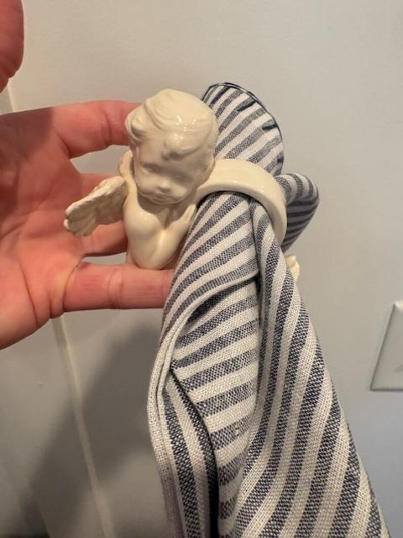 May include: A cream-colored ceramic napkin ring featuring a cherub with small wings. The cherub holds a blue and white striped napkin. The napkin ring is a decorative item for the dining table.