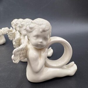 May include: A collection of ivory-colored cherub napkin rings. Each cherub is in a seated position with outstretched wings and a ring-shaped opening for a napkin. The cherubs are arranged in a row on a dark surface.