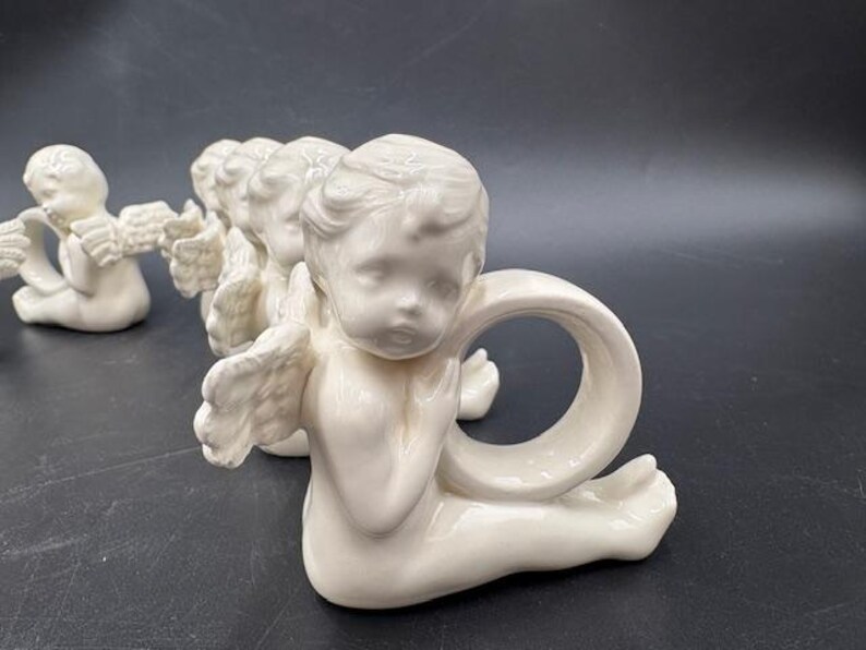 May include: A collection of ivory-colored cherub napkin rings. Each cherub is in a seated position with outstretched wings and a ring-shaped opening for a napkin. The cherubs are arranged in a row on a dark surface.