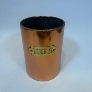 May include: A cylindrical copper container with a smooth, reflective finish. Attached to the front is a rectangular brass-colored metal plaque with rounded ends, featuring the word TOOLS embossed in capital letters with a rivet on each side.