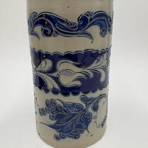 May include: A cylindrical stoneware vase featuring a light gray base with three horizontal bands of deep blue embossed floral and vine patterns. The top rim is finished with a scalloped blue edge, and the surface displays a smooth, glazed texture.