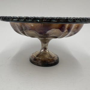 May include: A vintage silver-toned pedestal bowl with a scalloped rim. The bowl has a rounded, fluted body and a short, pedestal base. The metal has a tarnished appearance, with hints of purple and brown. This decorative bowl is suitable for serving or display.